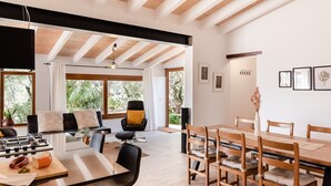 A modern kitchen with a central island, dining area with wooden chairs, and large windows offering a view of greenery. - Country House 'Ses Cases' with Mountain View, Private Pool and Wi-Fi (Manacor)