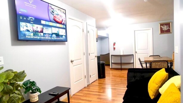 A living room with a flat-screen TV mounted on the wall, a black sofa with yellow cushions, a wooden coffee table, and a potted plant. - Lovely Loft Near NYC, EWR &
American Dream Mall (Newark)