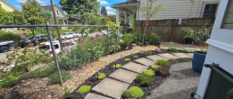 Un jardín residencial con un camino de piedra, diversas plantas y una cerca de madera.