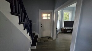 Una escalera conduce a una puerta con un pequeño ventanal, un televisor sobre un mueble, y vistas al exterior con vegetación.