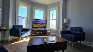 Una sala moderna con dos sillones azules, una mesa de centro de madera, un televisor de pantalla plana sobre un mueble de madera y ventanas grandes con marcos blancos.