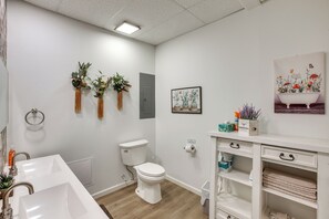 Een moderne badkamer met een witte wasbak, toilet en badkuip. Er staan drie potplanten op de muur, een kunstwerk met bloemen in een lijst en een wit kastje met lades.