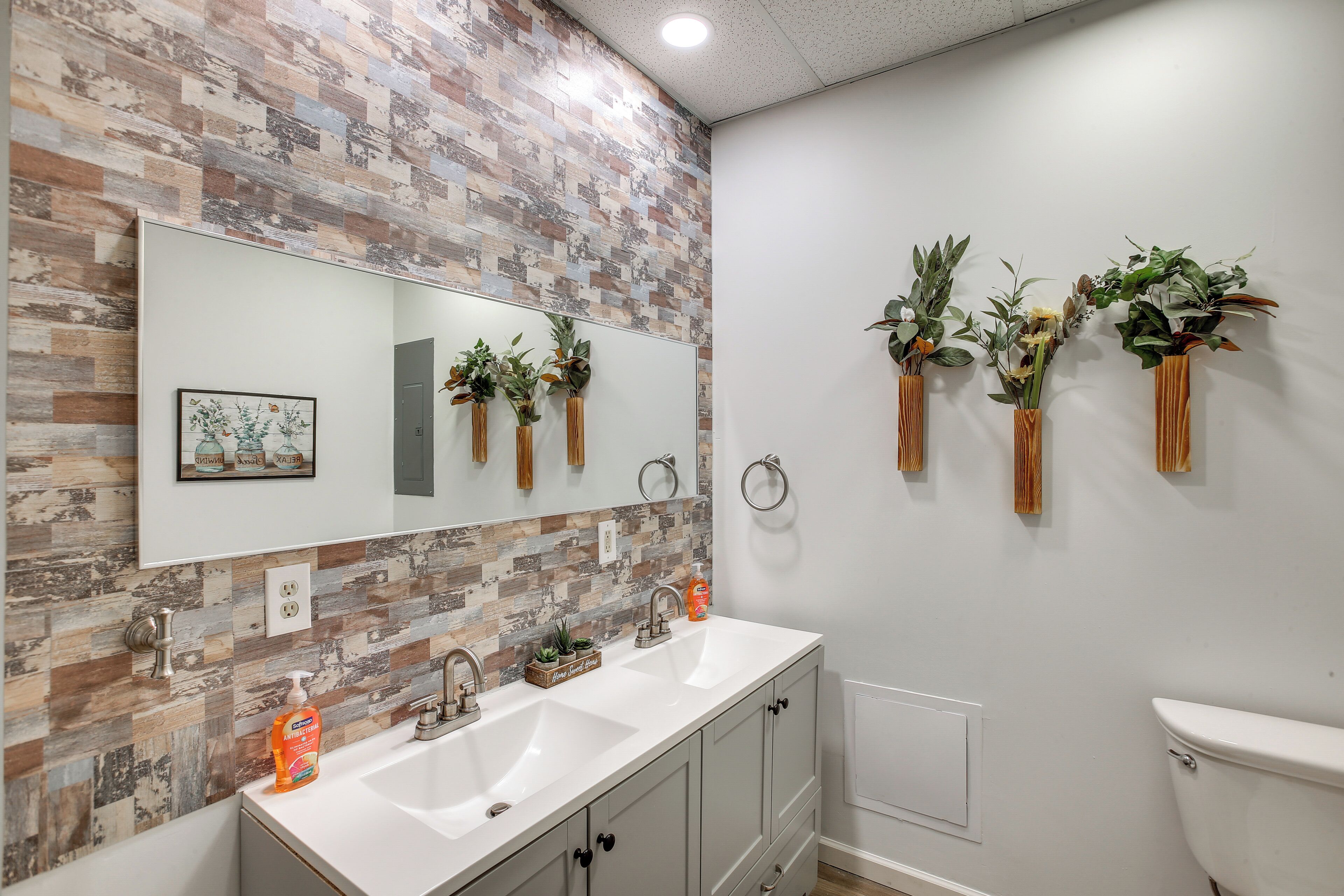 A bathroom with a white sink, two faucets, a mirror, and a toilet. There are three wooden vases with plants on the wall.
