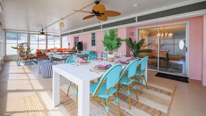 A spacious indoor area with a dining table set for four, surrounded by blue and white chairs. There are ceiling fans, a large window, and a door leading to another room. - Costal Escape: Heated Pool~Tiki Bar~Hot Tub~Fire Pit~Acarde~Pool Table (Bradenton)