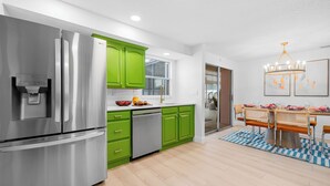 A modern kitchen with stainless steel appliances, green cabinets, and a dining area with a chandelier. - Costal Escape: Heated Pool~Tiki Bar~Hot Tub~Fire Pit~Acarde~Pool Table (Bradenton)