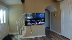 A spacious living area with a wooden floor, a staircase, and a mounted flat-screen TV displaying a streaming service interface. - 4Bd 2Ba 7 Person Sleeper-  King Bed (Somerdale)