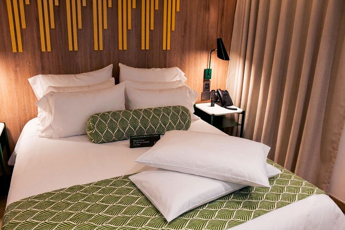 A hotel room with a bed, pillows, a nightstand with a lamp, and a curtain.