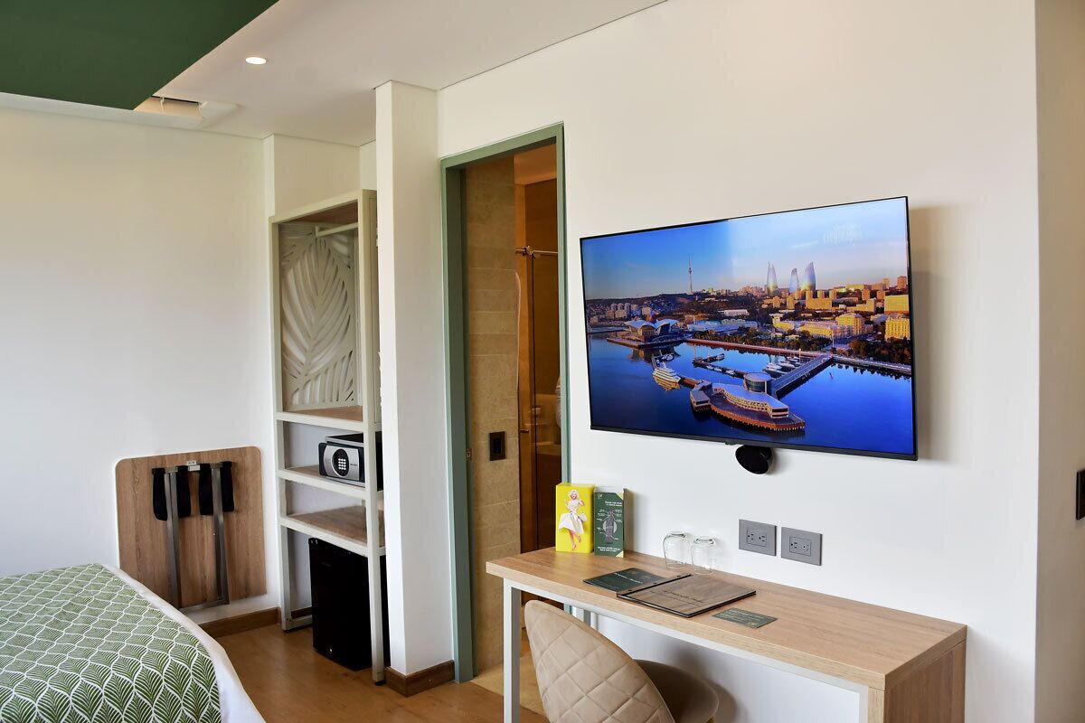 A hotel room with a bed, a desk with a chair, a television mounted on the wall, and a view of a cityscape.