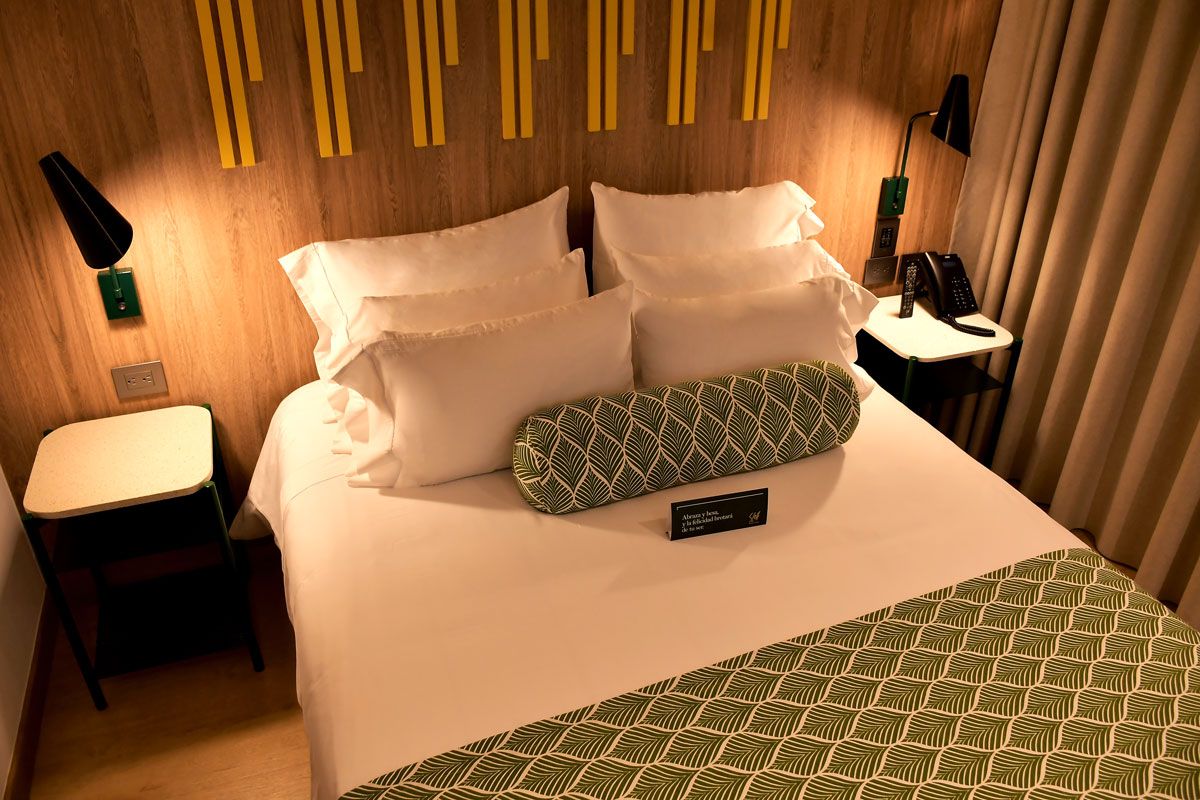 A hotel room with a bed, a bedside table with a lamp, a chair, and a nameplate on the bed.