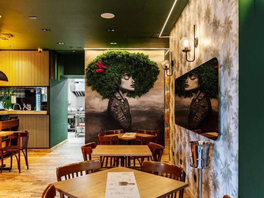 L’intérieur d’un restaurant avec des tables et des chaises en bois, un comptoir où est affiché le menu, et une fresque murale représentant le portrait d’une femme.