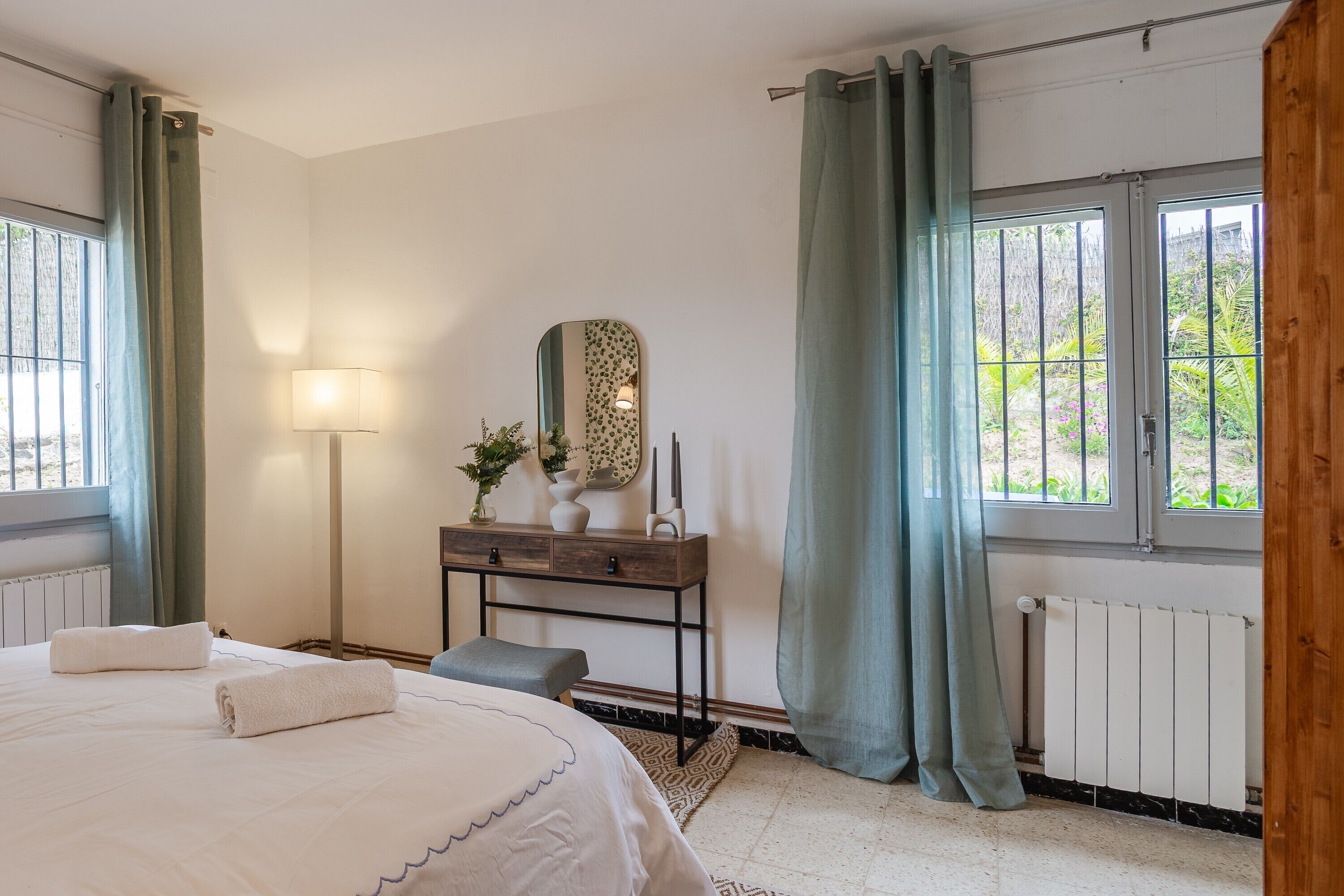 Een slaapkamer met een bed, nachtkastje, lamp, spiegel en uitzicht door een raam.