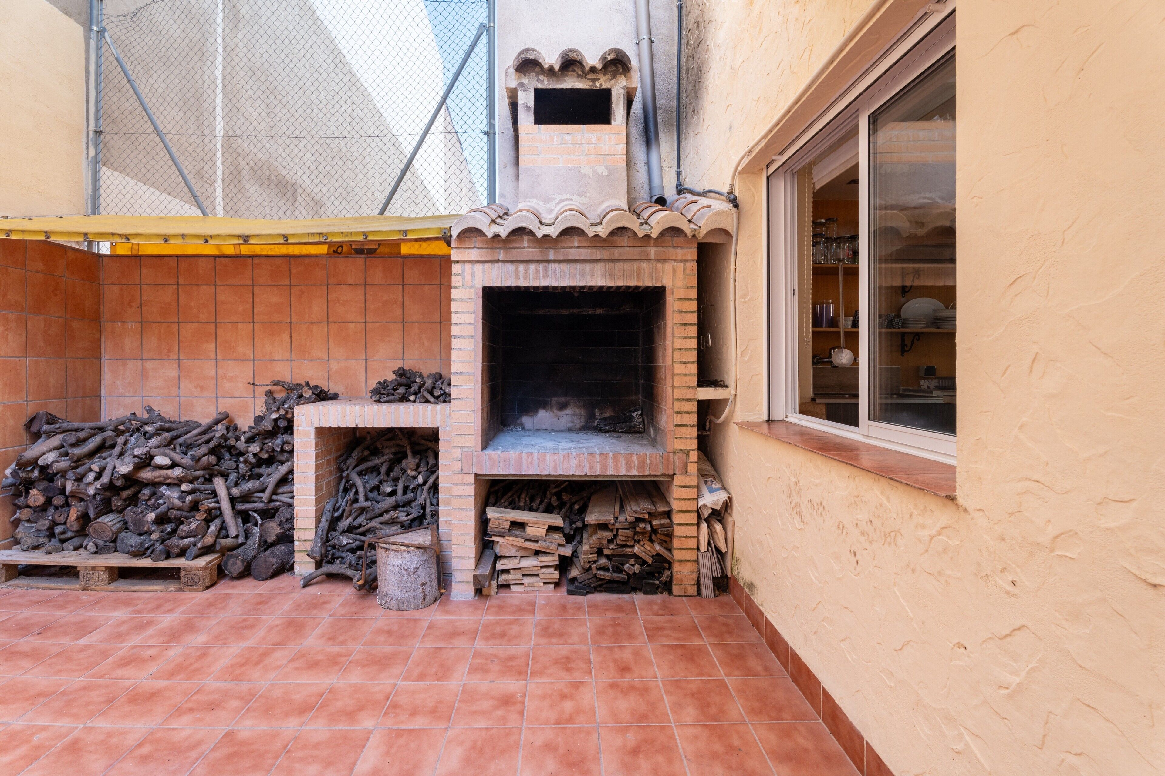 Ein gemauerter Kamin im Freien, eine geflieste Terrasse, ein Fenster mit Regalen und ein Holzstoß.