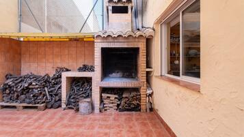 Ein gemauerter Kamin im Freien, eine geflieste Terrasse, ein Fenster mit Regalen und ein Holzstoß.