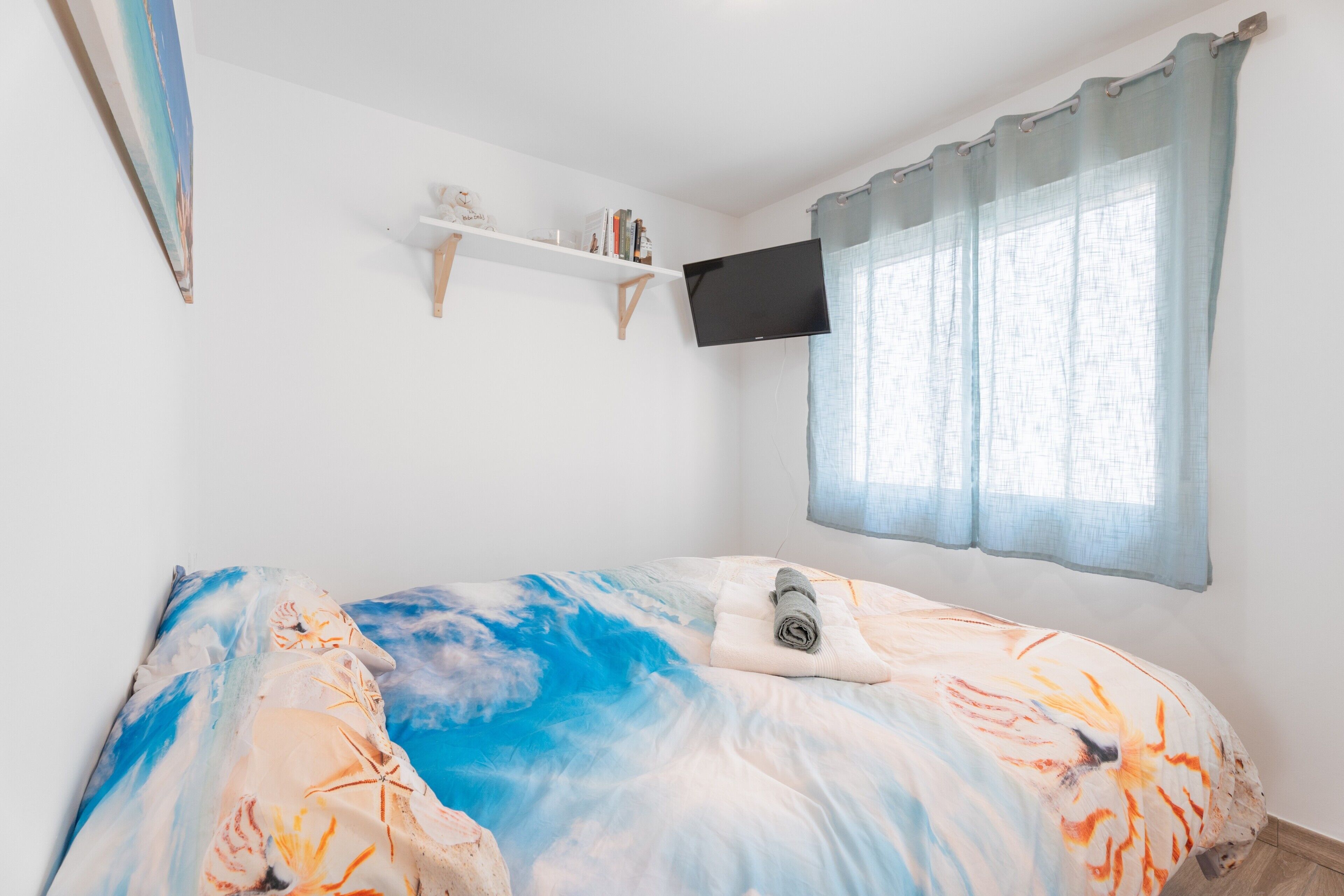 Un dormitorio con cama, televisor montado, ventana con cortinas y una estantería con libros.