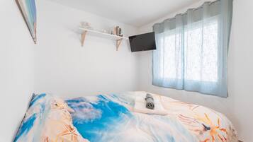 Un dormitorio con cama, televisor montado, ventana con cortinas y una estantería con libros.