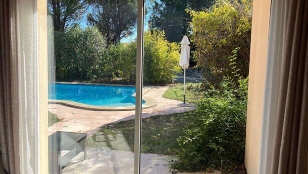 Vista attraverso una porta vetrata su un giardino con piscina, circondato da vegetazione e un'area patio.