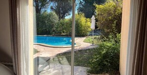 A view through a glass door of a backyard with a swimming pool, surrounded by greenery and a patio area.