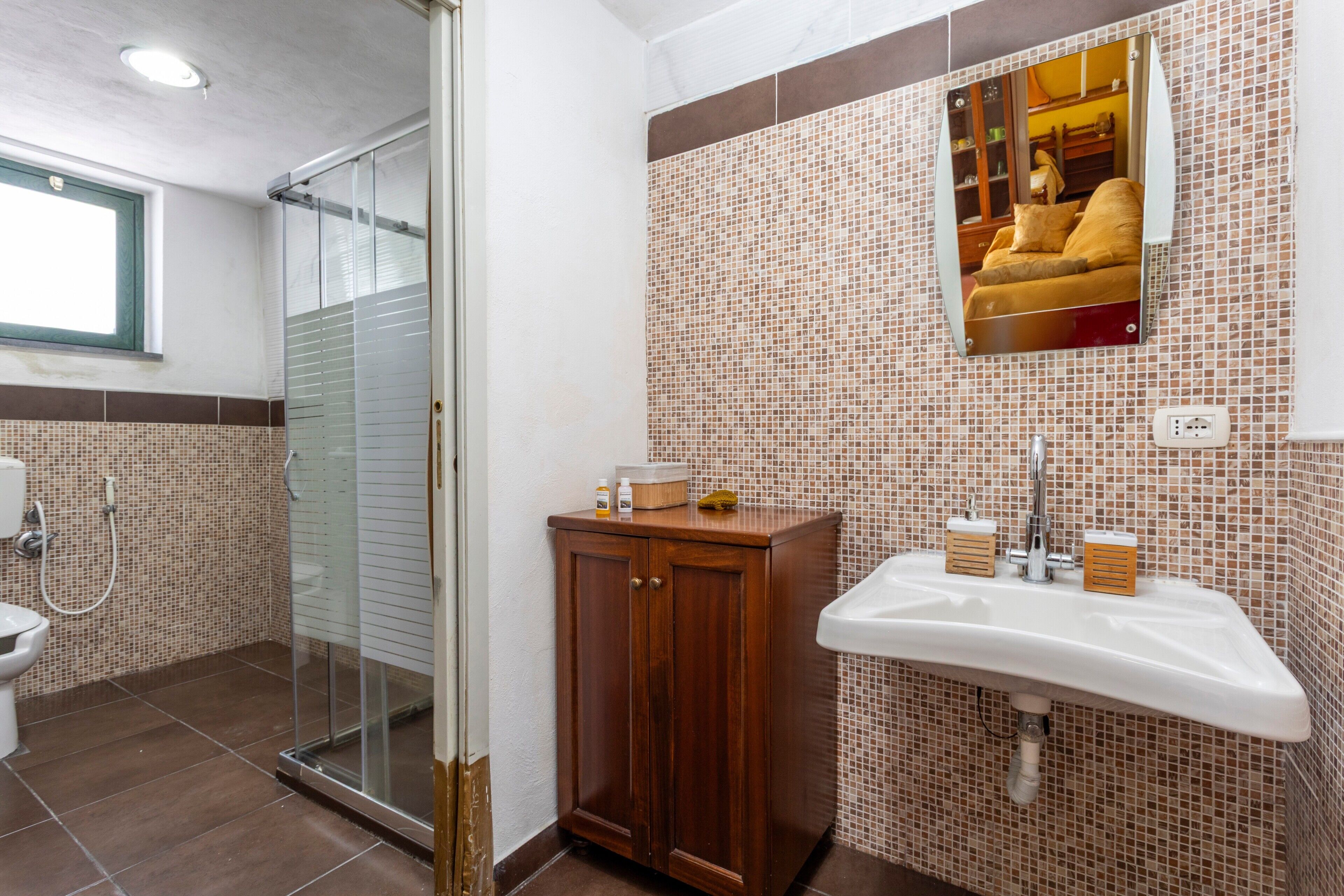 Une salle de bain dotée d’une douche à parois de verre, d’un meuble en bois, d’un lavabo avec distributeurs de savon et d’un miroir dans lequel se reflète un lit.