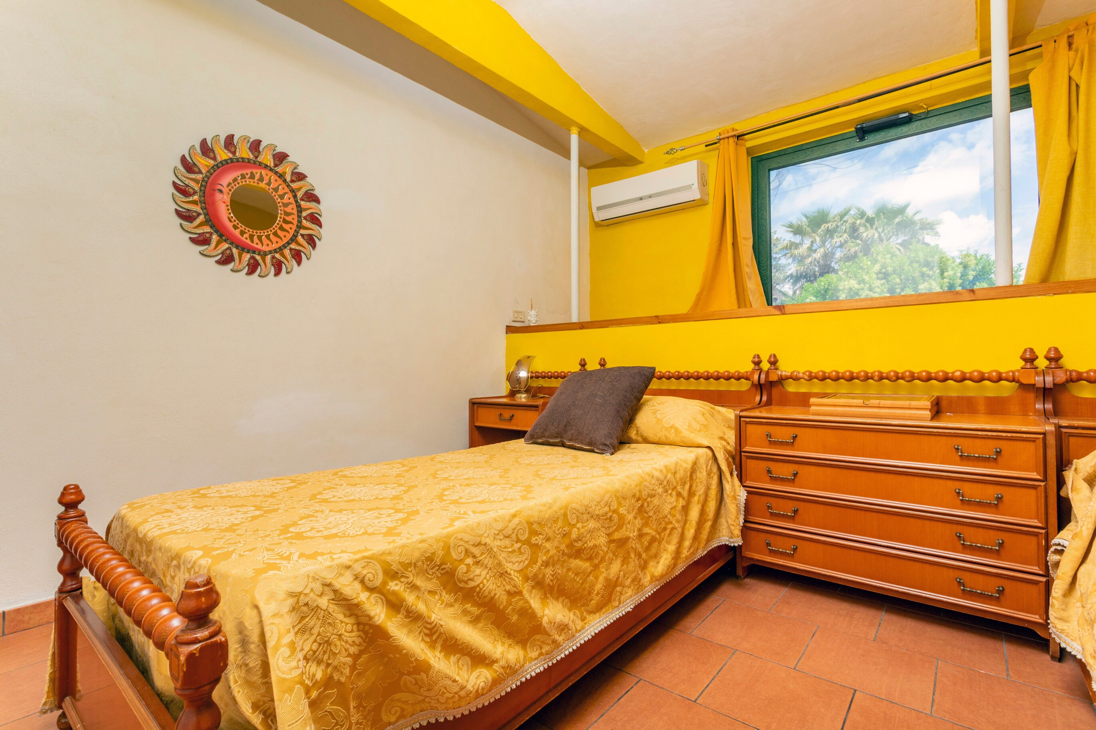 Een slaapkamer met een bed, een houten dressoir, een nachtkastje, een wanddecoratie en een raam met gordijnen.