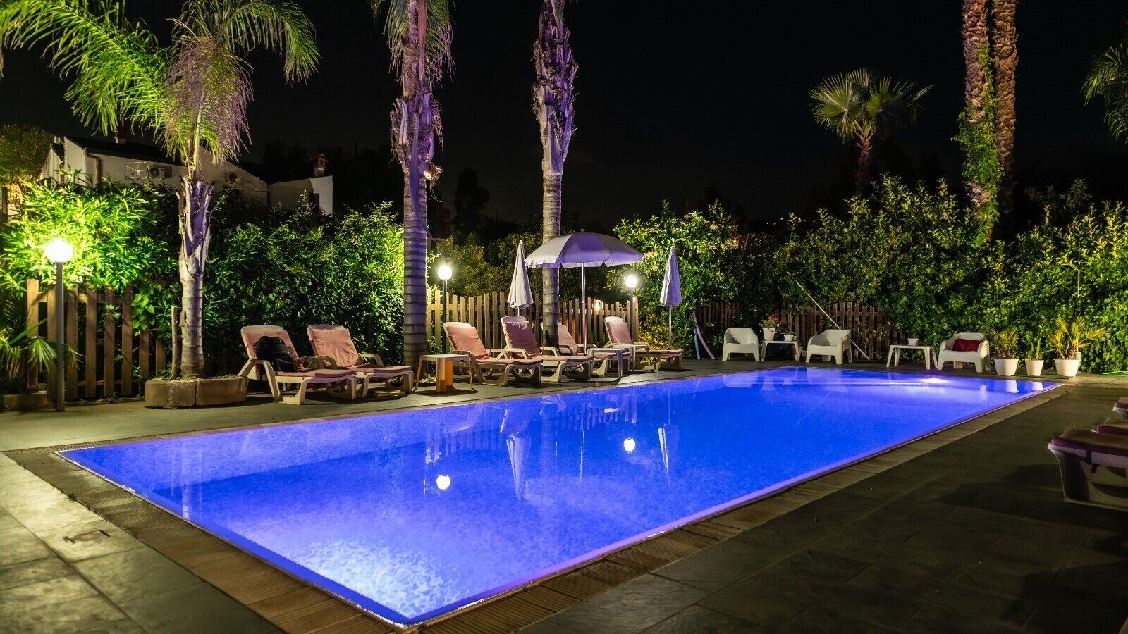 Una piscina nocturna con sillones de descanso y sombrillas, rodeada de palmeras y una valla de madera.