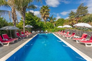 A swimming pool with red lounge chairs and umbrellas, surrounded by palm trees and greenery.