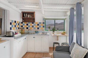 Une cuisine compacte avec des meubles blancs, un pare-feu en carrelage, une fenêtre offrant une belle vue et un canapé gris agrémenté d’un coussin.