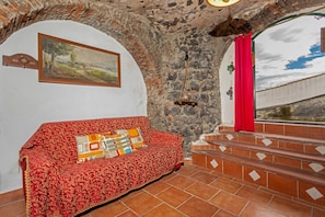 Une pièce avec un canapé rouge à motifs, un tableau au mur, des murs en pierre et une vue par la fenêtre.