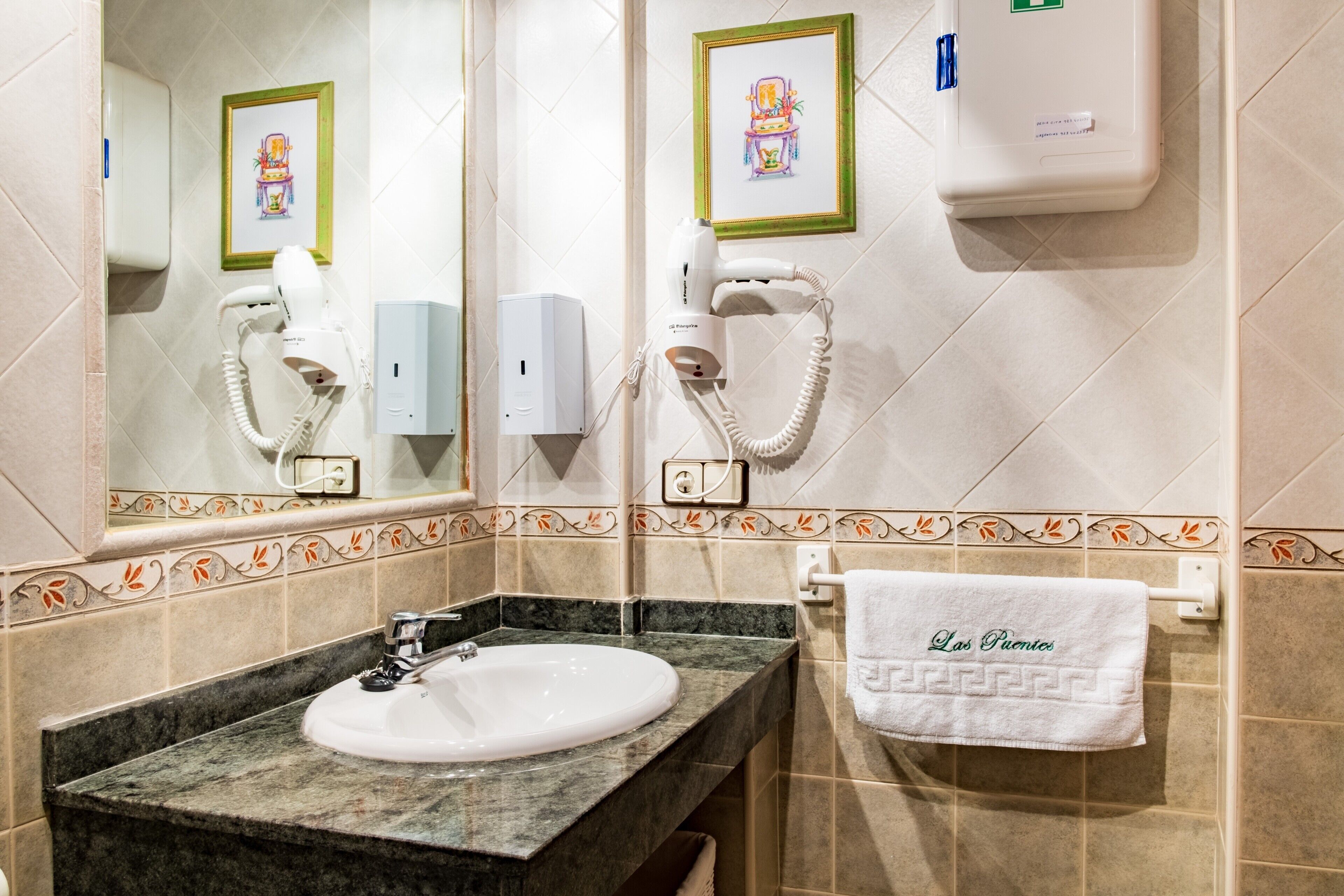 Un baño con lavamanos, dos secadores de pelo, obra de arte enmarcadas y un perchero con toalla.