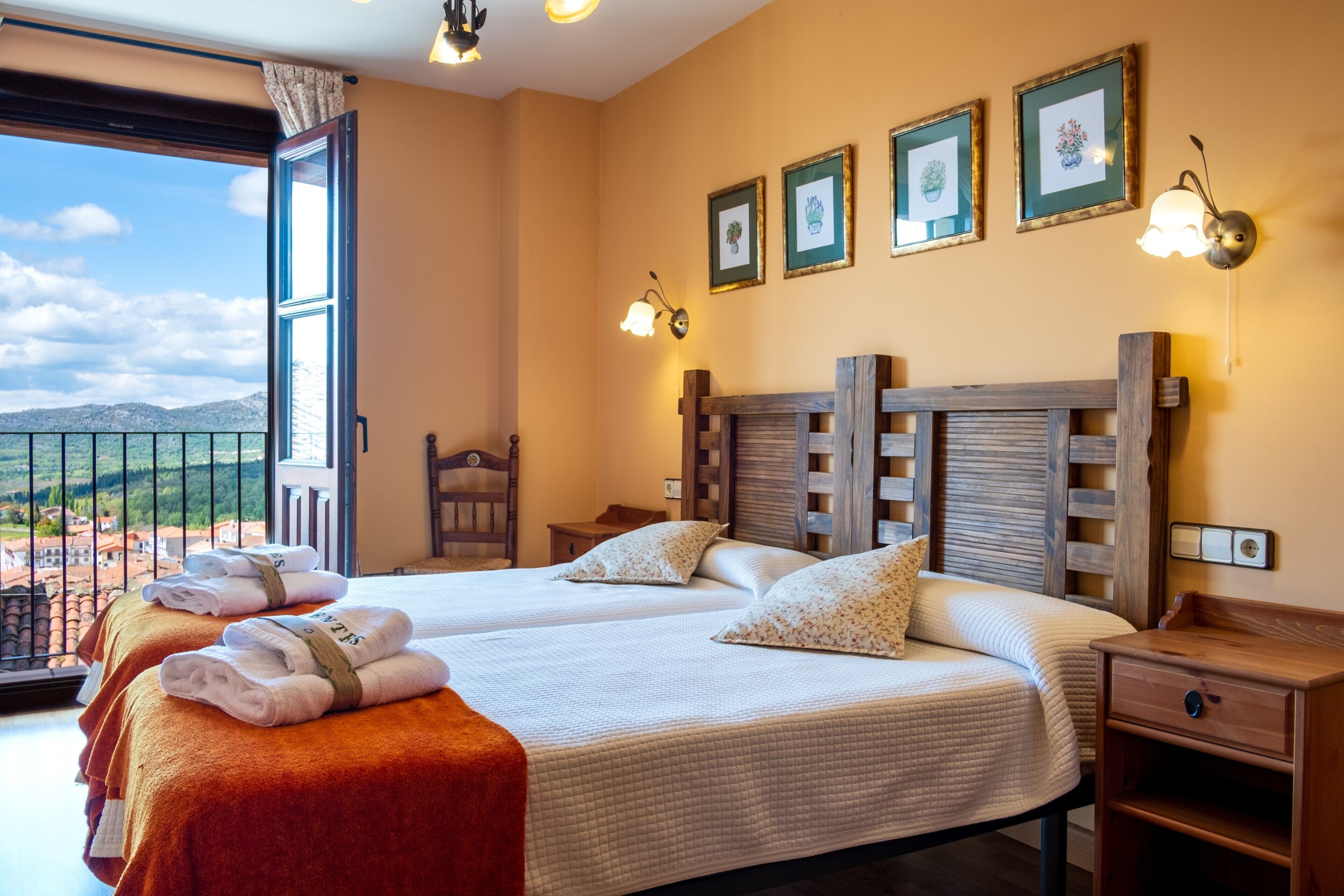 A hotel room with two beds, a wooden headboard, a nightstand, a chair, and a balcony with a view of the countryside.