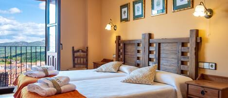 A hotel room with two beds, a wooden headboard, a nightstand, a chair, and a balcony with a view of the countryside.