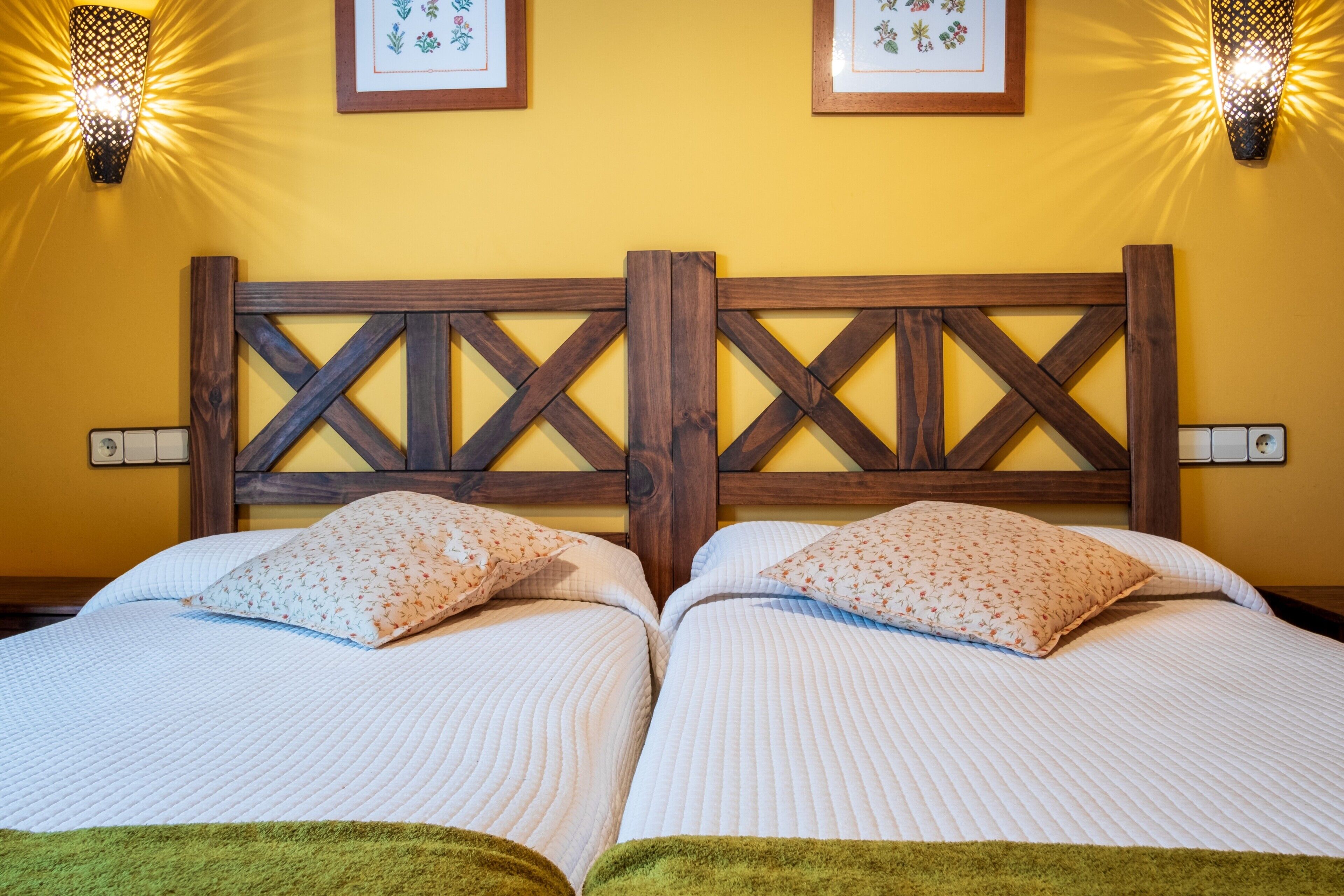 Een tweepersoonsbed met een houten hoofdbord, twee kussens, een groene plaid en twee wandlampen.