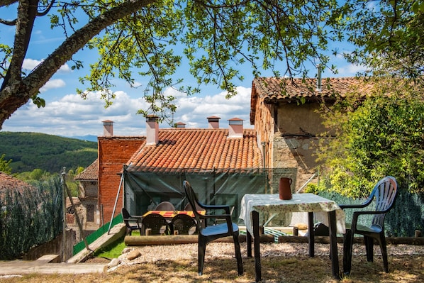 A rustic outdoor dining area with a table and chairs, surrounded by a brick building with a tiled roof.