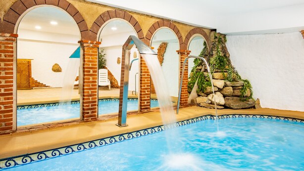 Una piscina interior con cascada, arcos de ladrillo y una pared de piedra con vegetación.