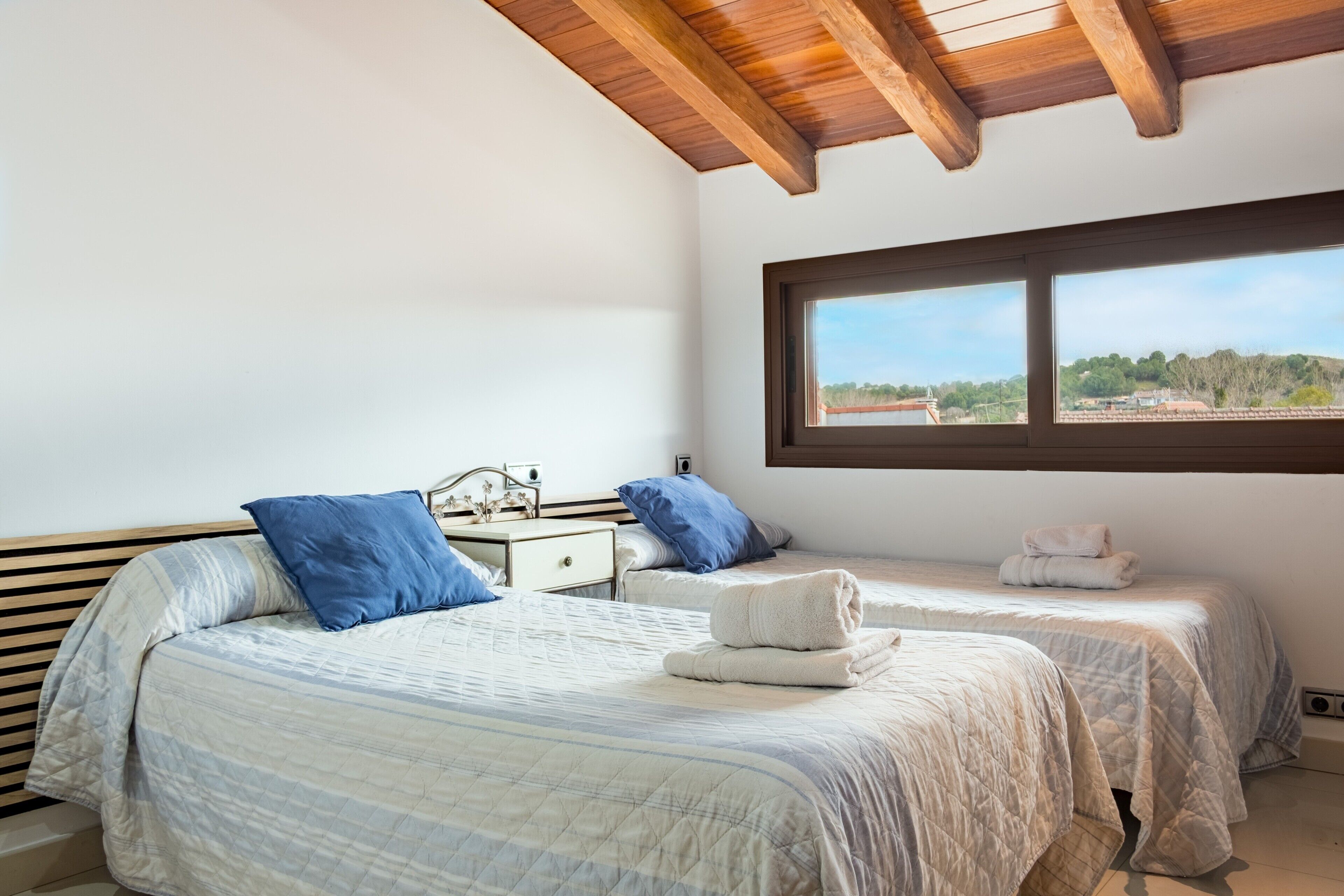 A bedroom with two beds, a window with a view, wooden beams on the ceiling, and a nightstand with a lamp.
