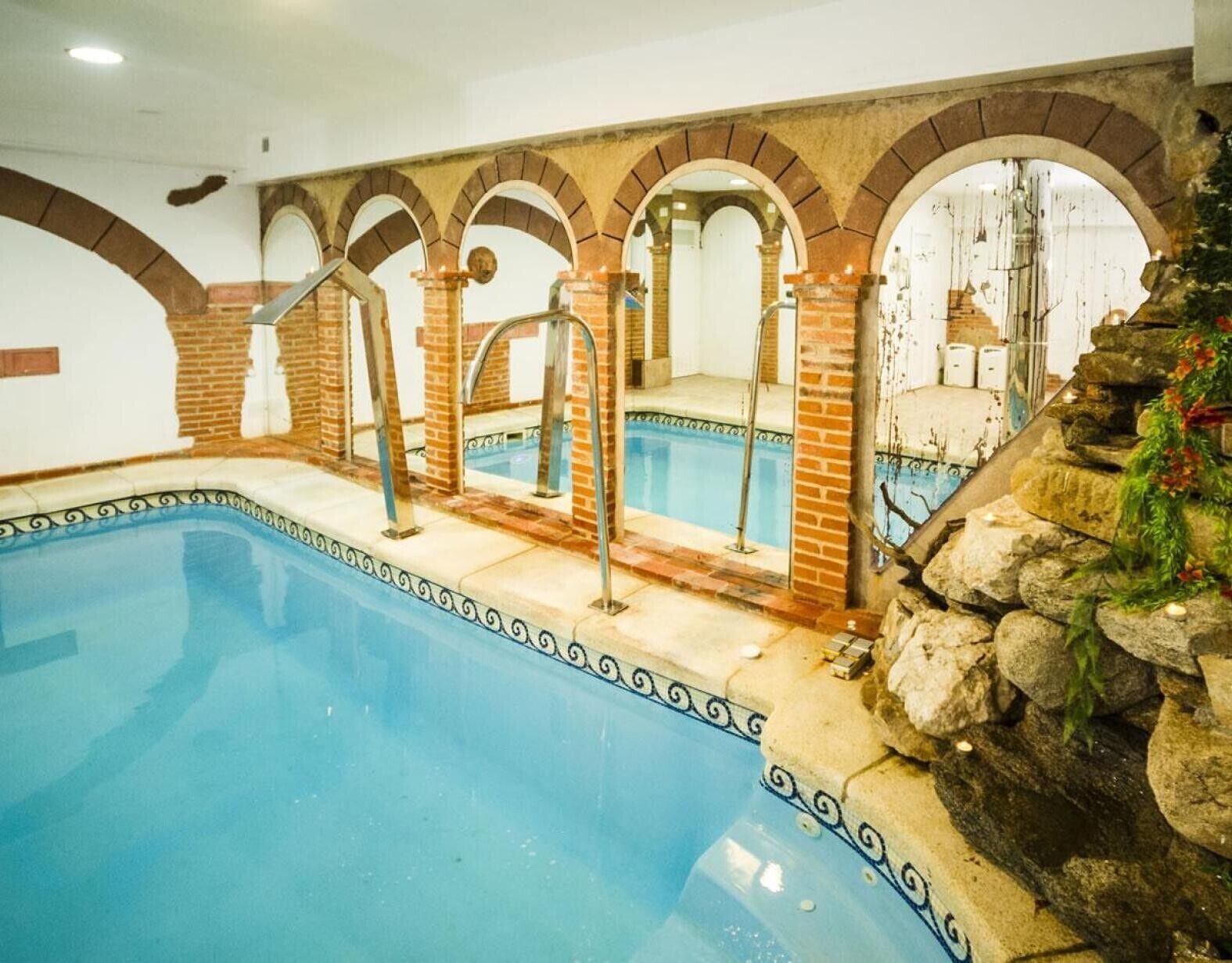 Una piscina interior con borde de azulejos, arcos de ladrillo y una pared de piedra con vegetación.