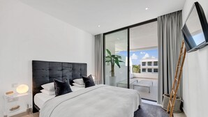 A modern bedroom with a large bed, a glass door leading to a balcony, a mounted TV, and a clear nightstand. - Luxury 2-Bed Apartment with Stunning 81 m2 Terrace & Lagoon-Style Pool (Estepona)