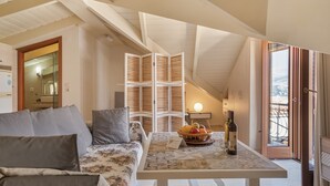 Quarto aconchegante com sofá, mesa com uma cesta de frutas e uma garrafa de vinho, e vista através de uma porta aberta.