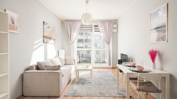A modern living room with a sofa, a dining table, and a chair. There is a window with curtains, a hanging light fixture, and a vase with flowers on the table. - Warszawa-Włochy Serene Apartment (Warszawa)