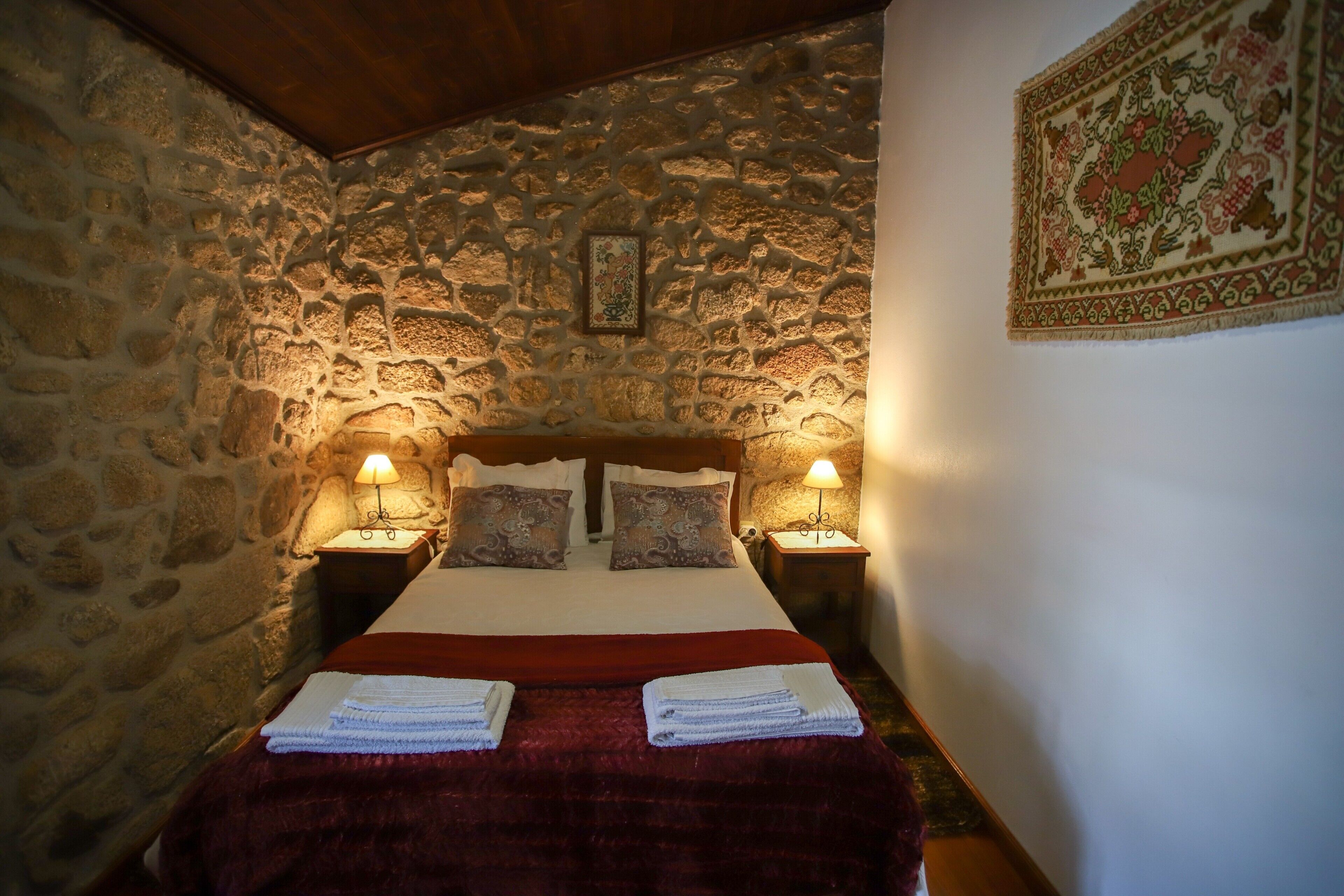 Un dormitorio de paredes de piedra con una cama, dos lámparas de noche y un tapiz decorativo.
