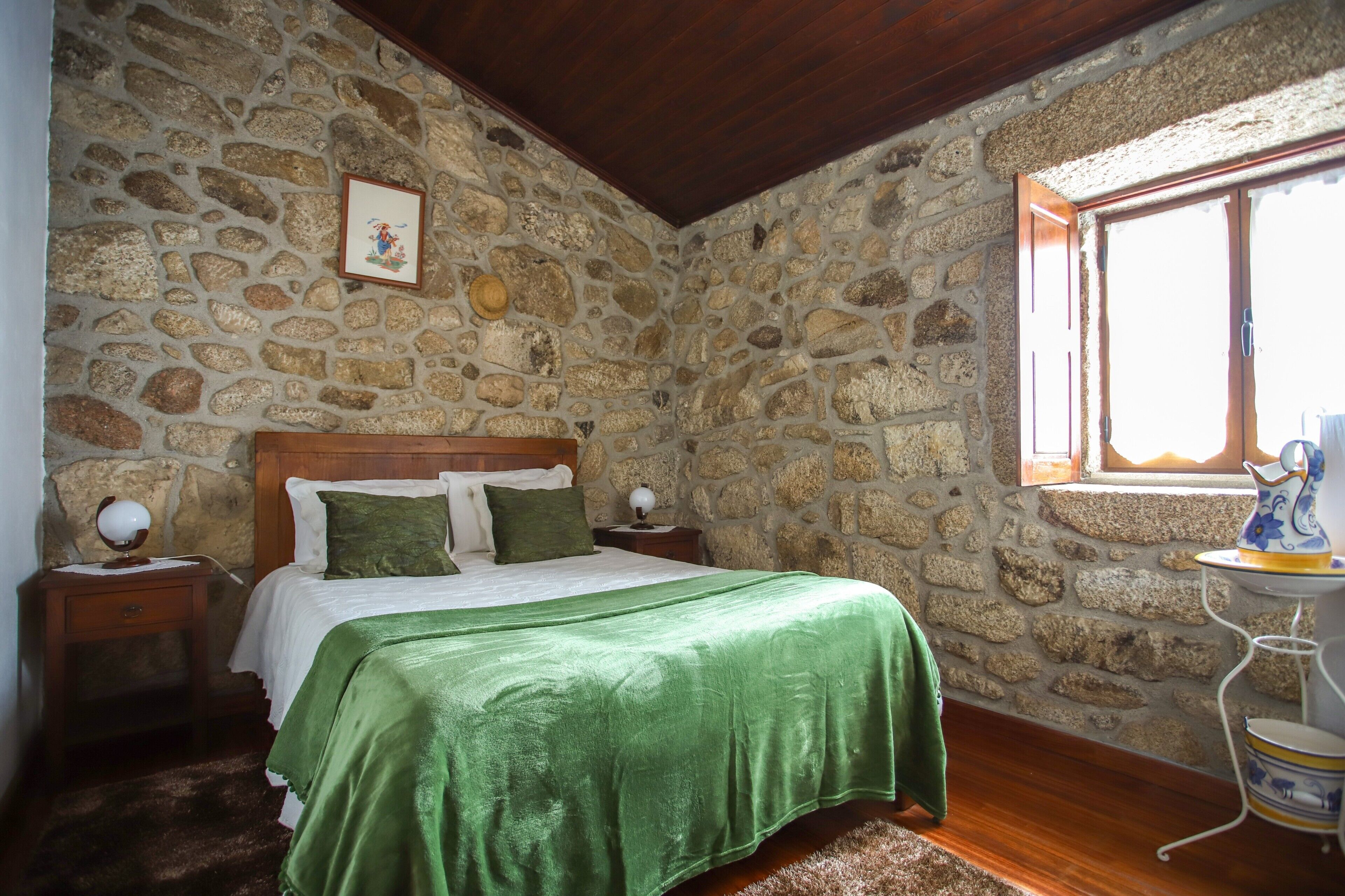 A bedroom with a stone wall, a wooden ceiling, a bed with green bedding, a nightstand with a lamp, and a window with a view.