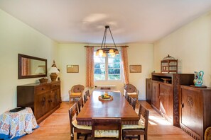Ein Esszimmer mit einem langen Holztisch, Korbstühlen, einer Chandelierleuchtre und einem Fenster mit Vorhängen.