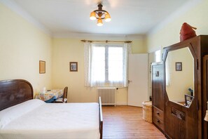 Ein Schlafzimmer mit Bett, Kommode, Stuhl, Tisch, einem Fenster mit Vorhängen, einem Heizkörper und einer Deckenleuchte.