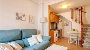 A living room with a blue sofa, a white table, and a kitchen area with wooden cabinets and a washing machine. - Bungalow 'Casa De Verónica' with Private Terrace, Wi-Fi and Air Conditioning (Santa Pola)