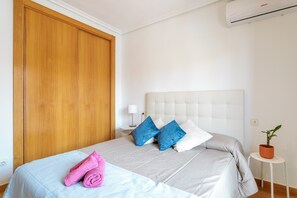 A bedroom with a bed, a wooden wardrobe, a nightstand, a lamp, and a potted plant.