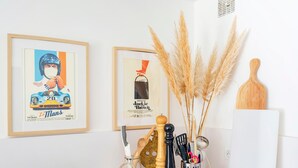 Una pared con dos carteles enmarcados, un cuenco de madera, un recipiente metálico con utensilios de cocina y un florero con plantas secas.