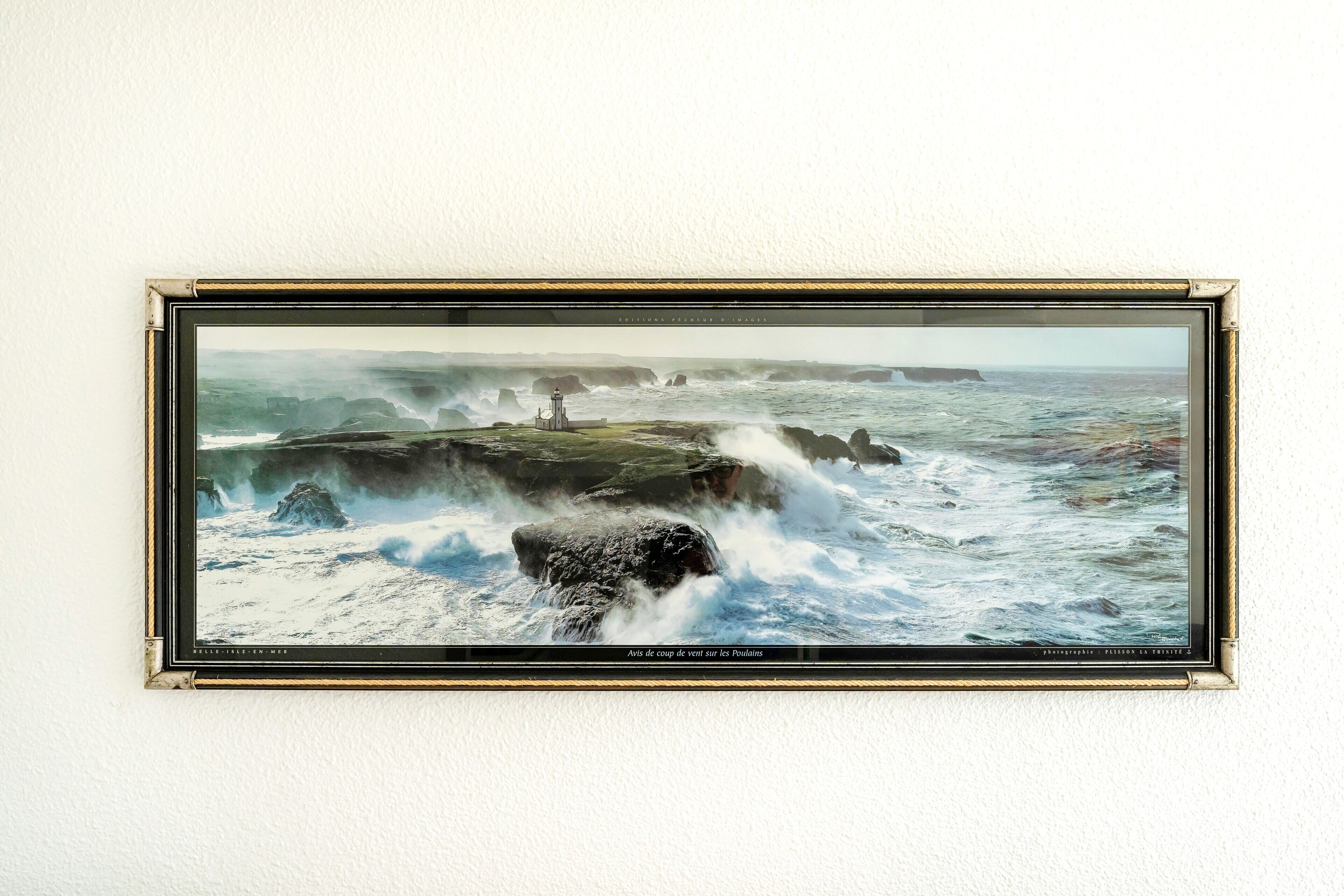 Un tableau encadré représentant un paysage côtier, avec des vagues se brisant contre des rochers et un phare.