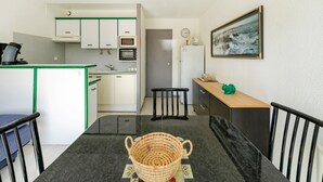 Une cuisine compacte avec des meubles blancs, un four à micro-ondes et un réfrigérateur. On y trouve une table à manger avec des chaises et un panier en osier au centre.