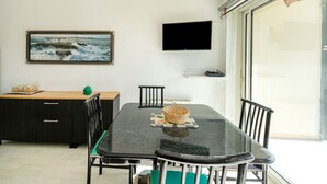 Un coin repas avec une table en verre, des chaises, un téléviseur fixé au mur et un tableau représentant un paysage côtier.