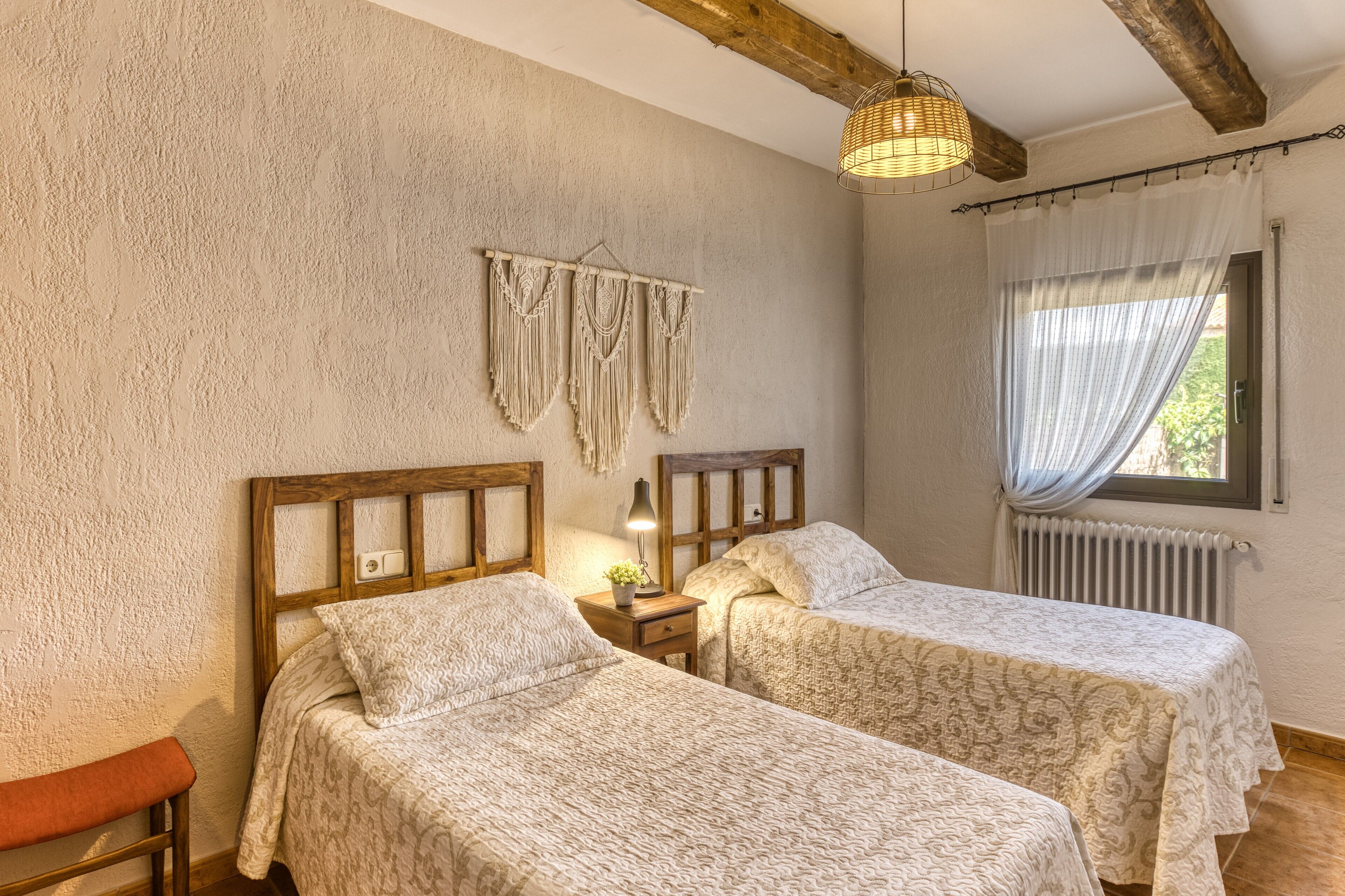 A room with two beds, a wooden bench, a macrame wall hanging, a lamp, and a window with curtains.