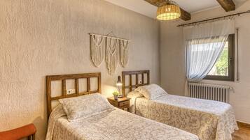 A room with two beds, a wooden bench, a macrame wall hanging, a lamp, and a window with curtains.